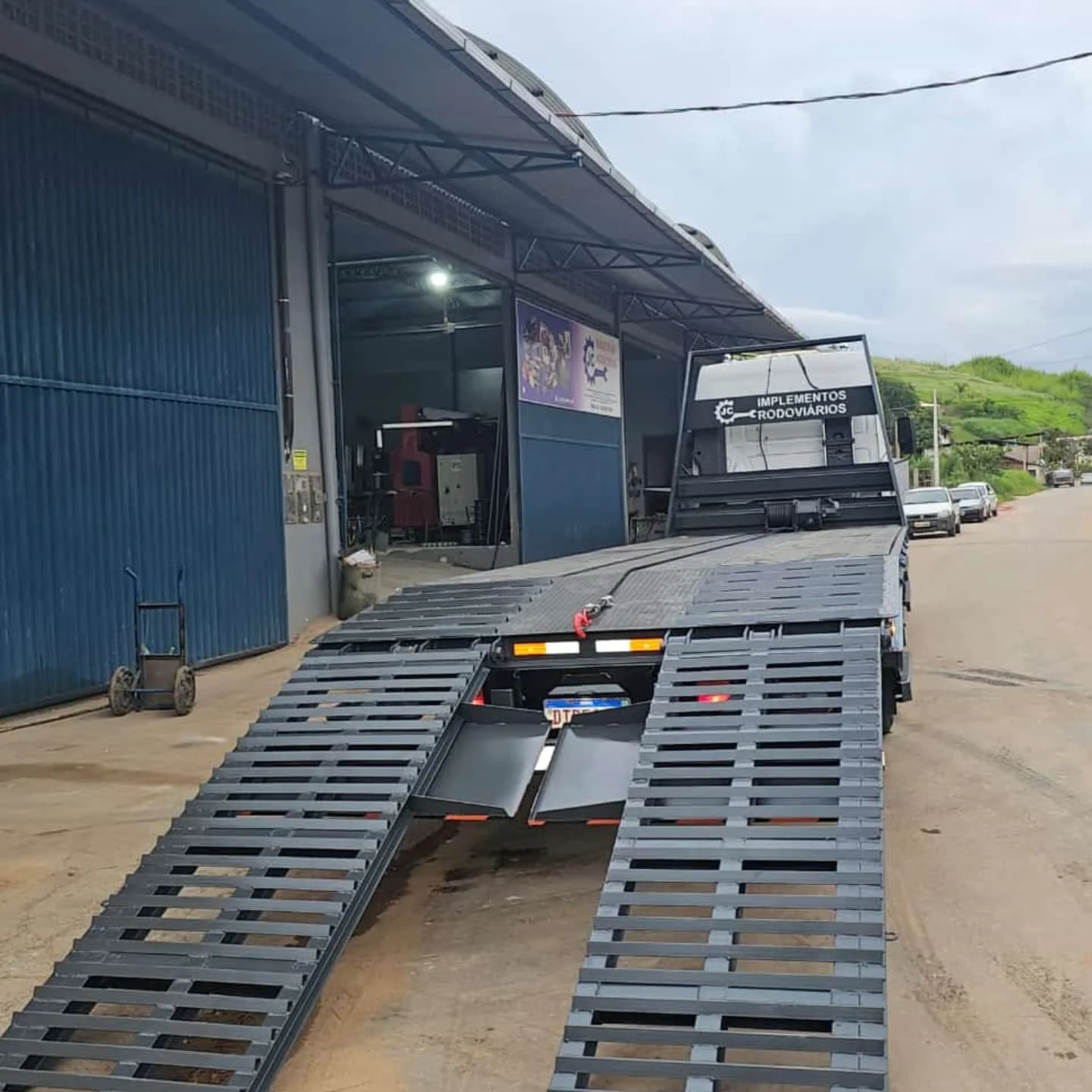 Caminhão com plataforma sendo carregado na entrada da oficina da JC, com maquinário industrial visível ao fundo